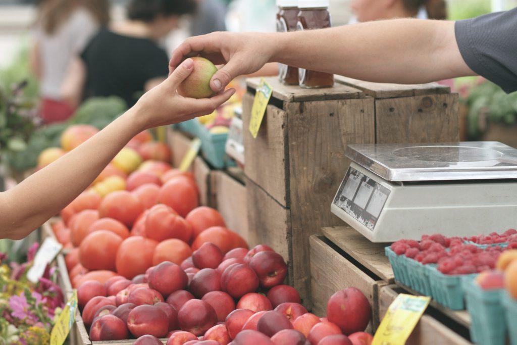 A photo in which one person hands an apple to another person at a fruit stand.